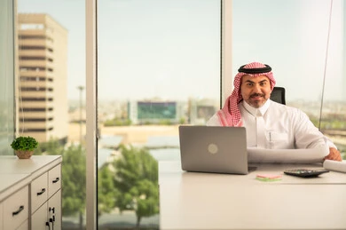 Reviewing project plans, gestures of integration and focus, providing a comfortable and suitable work environment, using a laptop to complete tasks professionally, a Saudi Arabian Gulf man wearing a traditional thobe and shemagh sitting in an office working on a laptop, employment in Saudi offices and companies