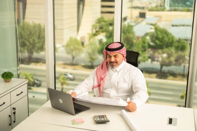 تدقيق مخططات المشاريع، إيماءات الاندماج والتركيز، توفير بيئة عمل مريحة ومناسبة، استخدام جهاز الحاسوب المحمول لإنجاز المهام باحترافية، رجل عربي خليجي سعودي يرتدي الثوب والشماغ التقليدي يجلس في المكتب يقوم بالعمل عبر اللابتوب، التوظيف في المكاتب والشركات السعودية