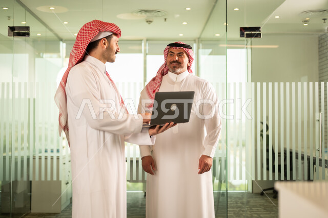 استخدام الحاسب والتقنية في مجال الاعمال، بيئة العمل المريحة في الشركات السعودية، الحوار و النقاش بين الزملاء، تبادل الخبرات والمعلومات لتحقيق الاهداف, موظفان عربيان خليجيان سعوديان يقومان بالعمل ومشاركة الأفكار داخل مقر الشركة