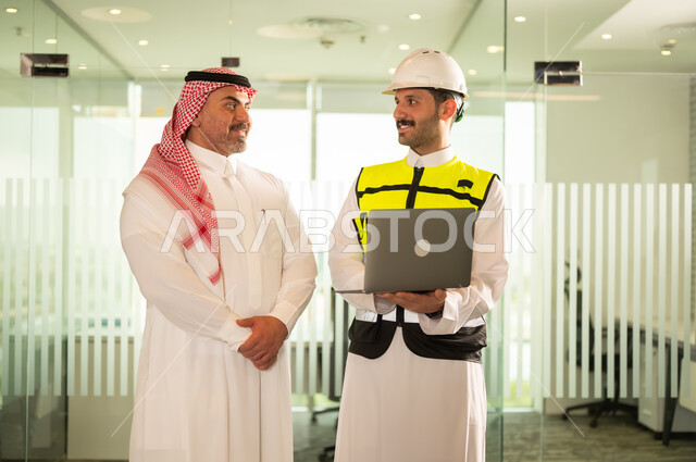 الشركات المختصة بورشات البناء والمقاولات، تصميم وتخطيط المخططات المعمارية عبر الحاسوب المحمول، متابعة تنفيذ الخطط الهندسية، رجل أعمال عربي خليجي سعودي يرتدي الشماغ ومهندس يرتدي الخوذة وسترة الحماية يقومان بالمناقشة وتبادل الخبرات، استخدام جهاز تقني حديث