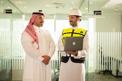 الشركات المختصة بورشات البناء والمقاولات، تصميم وتخطيط المخططات المعمارية عبر الحاسوب المحمول، متابعة تنفيذ الخطط الهندسية، رجل أعمال عربي خليجي سعودي يرتدي الشماغ ومهندس يرتدي الخوذة وسترة الحماية يقومان بالمناقشة وتبادل الخبرات، استخدام جهاز تقني حديث