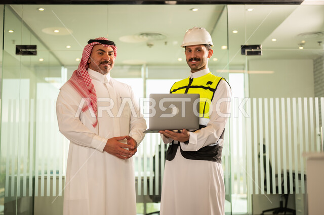النظر إلى الكاميرا بتعابير السرور، الشركات المختصة بورشات البناء والمقاولات، تصميم وتخطيط المخططات المعمارية عبر الحاسوب المحمول، رجل أعمال عربي خليجي سعودي يرتدي الشماغ ومهندس يرتدي الخوذة وسترة الحماية يقومان بالمناقشة وتبادل الخبرات، استخدام جهاز تقني حديث