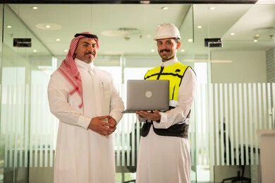 Looking at the camera with happy expressions, companies specialized in construction and contracting workshops, designing and planning architectural plans via laptop, a Saudi Arabian Gulf businessman wearing a shemagh and an engineer wearing a helmet and a protective vest discussing and exchanging experiences, using a modern technical device