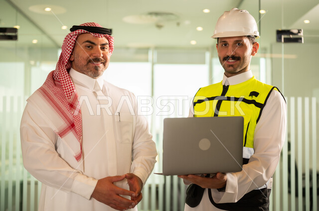 النظر إلى الكاميرا بتعابير السرور، الشركات المختصة بورشات البناء والمقاولات، تصميم وتخطيط المخططات المعمارية عبر الحاسوب المحمول، رجل أعمال عربي خليجي سعودي يرتدي الشماغ ومهندس يرتدي الخوذة وسترة الحماية يقومان بالمناقشة وتبادل الخبرات، استخدام جهاز تقني حديث