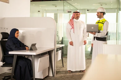 الشركات المختصة بورشات البناء والمقاولات، تصميم وتخطيط المخططات المعمارية عبر الحاسوب المحمول، متابعة تنفيذ الخطط الهندسية، رجل أعمال عربي خليجي سعودي يرتدي الشماغ ومهندس يرتدي الخوذة وسترة الحماية يقومان بالمناقشة وتبادل الخبرات، استخدام جهاز تقني حديث