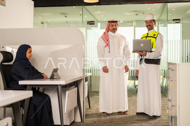 الشركات المختصة بورشات البناء والمقاولات، تصميم وتخطيط المخططات المعمارية عبر الحاسوب المحمول، متابعة تنفيذ الخطط الهندسية، رجل أعمال عربي خليجي سعودي يرتدي الشماغ ومهندس يرتدي الخوذة وسترة الحماية يقومان بالمناقشة وتبادل الخبرات، استخدام جهاز تقني حديث