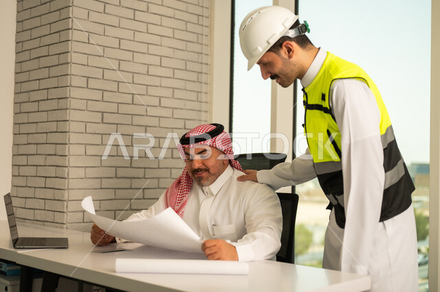 الشركات المختصة بورشات البناء والمقاولات، التخطيط ووضع اللمسات الأخيرة على المخططات المعمارية، رجل أعمال عربي خليجي سعودي يرتدي الشماغ ومهندس يرتدي الخوذة وسترة الحماية يعملان على مناقشة مخطط الإنشاء وتحديد القياسات بدقة، متابعة تنفيذ الخطط الهندسية الموضوعة مسبقا