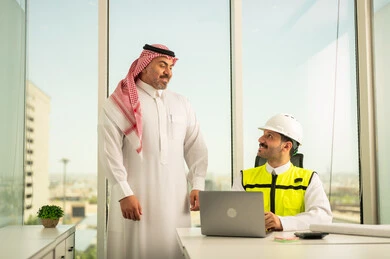 Companies specializing in construction and contracting workshops, designing and planning architectural plans via laptop, following up on the implementation of engineering plans, a Saudi Arabian Gulf businessman wearing a shemagh and an engineer wearing a helmet and a protective vest discussing and exchanging experiences, using a modern technical device