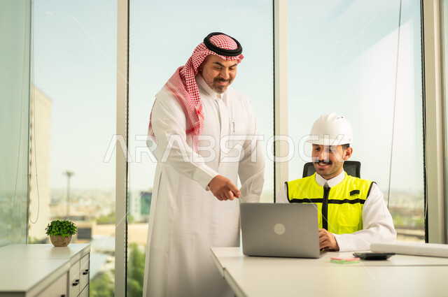 تصميم وتخطيط المخططات المعمارية عبر الحاسوب المحمول، متابعة تنفيذ الخطط الهندسية، الشركات المختصة بورشات البناء والمقاولات، رجل أعمال عربي خليجي سعودي يرتدي الشماغ ومهندس يرتدي الخوذة وسترة الحماية يقومان بالمناقشة وتبادل الخبرات، استخدام جهاز تقني حديث