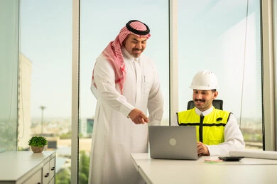 تصميم وتخطيط المخططات المعمارية عبر الحاسوب المحمول، متابعة تنفيذ الخطط الهندسية، الشركات المختصة بورشات البناء والمقاولات، رجل أعمال عربي خليجي سعودي يرتدي الشماغ ومهندس يرتدي الخوذة وسترة الحماية يقومان بالمناقشة وتبادل الخبرات، استخدام جهاز تقني حديث