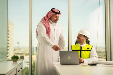 تصميم وتخطيط المخططات المعمارية عبر الحاسوب المحمول، متابعة تنفيذ الخطط الهندسية، الشركات المختصة بورشات البناء والمقاولات، رجل أعمال عربي خليجي سعودي يرتدي الشماغ ومهندس يرتدي الخوذة وسترة الحماية يقومان بالمناقشة وتبادل الخبرات، استخدام جهاز تقني حديث