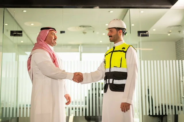 إيماءات السعادة والسرور، الشركات المختصة بورشات البناء والمقاولات، تصميم وتخطيط المخططات المعمارية، رجل أعمال عربي خليجي سعودي يرتدي الشماغ والثوب التقليدي يقوم بمصافحة مهندس يرتدي الخوذة وسترة الحماية داخل المكتب، عقد اتفاقيات عمل جديدة