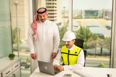 الشركات المختصة بورشات البناء والمقاولات، تصميم وتخطيط المخططات المعمارية عبر الحاسوب المحمول، متابعة تنفيذ الخطط الهندسية، رجل أعمال عربي خليجي سعودي يرتدي الشماغ ومهندس يرتدي الخوذة وسترة الحماية يقومان بالمناقشة وتبادل الخبرات، استخدام جهاز تقني حديث