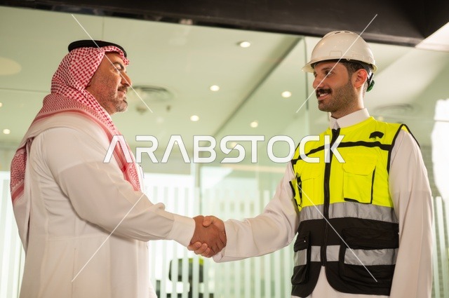عقد اتفاقيات عمل جديدة ، إيماءات السعادة والسرور، الشركات المختصة بورشات البناء والمقاولات، تصميم وتخطيط المخططات المعمارية، رجل أعمال عربي خليجي سعودي يرتدي الشماغ والثوب التقليدي يقوم بمصافحة مهندس يرتدي الخوذة وسترة الحماية داخل المكتب،