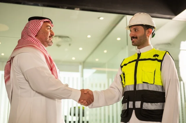 عقد اتفاقيات عمل جديدة ، إيماءات السعادة والسرور، الشركات المختصة بورشات البناء والمقاولات، تصميم وتخطيط المخططات المعمارية، رجل أعمال عربي خليجي سعودي يرتدي الشماغ والثوب التقليدي يقوم بمصافحة مهندس يرتدي الخوذة وسترة الحماية داخل المكتب،