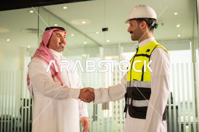 إيماءات السعادة والسرور، الشركات المختصة بورشات البناء والمقاولات، تصميم وتخطيط المخططات المعمارية، رجل أعمال عربي خليجي سعودي يرتدي الشماغ والثوب التقليدي يقوم بمصافحة مهندس يرتدي الخوذة وسترة الحماية داخل المكتب، عقد اتفاقيات عمل جديدة