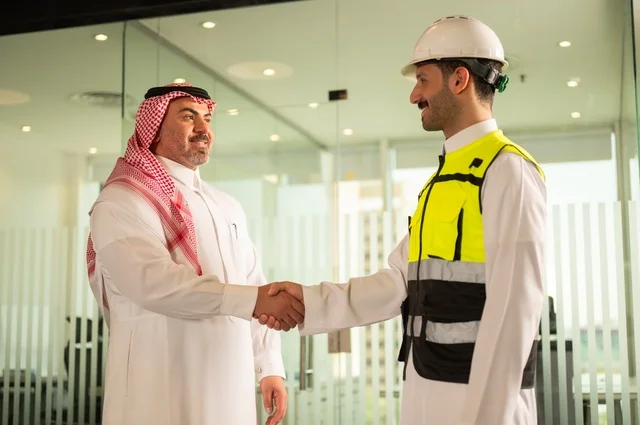 إيماءات السعادة والسرور، الشركات المختصة بورشات البناء والمقاولات، تصميم وتخطيط المخططات المعمارية، رجل أعمال عربي خليجي سعودي يرتدي الشماغ والثوب التقليدي يقوم بمصافحة مهندس يرتدي الخوذة وسترة الحماية داخل المكتب، عقد اتفاقيات عمل جديدة
