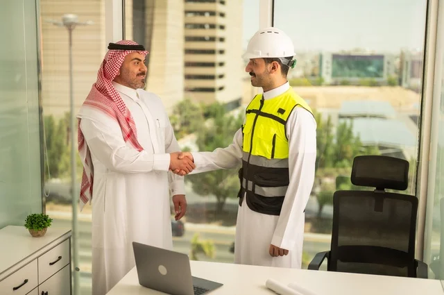 إيماءات السعادة والسرور، الشركات المختصة بورشات البناء والمقاولات، تصميم وتخطيط المخططات المعمارية، رجل أعمال عربي خليجي سعودي يرتدي الشماغ والثوب التقليدي يقوم بمصافحة مهندس يرتدي الخوذة وسترة الحماية داخل المكتب، عقد اتفاقيات عمل جديدة