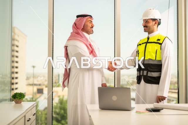 عقد اتفاقيات عمل جديدة ، إيماءات السعادة والسرور، الشركات المختصة بورشات البناء والمقاولات، تصميم وتخطيط المخططات المعمارية، رجل أعمال عربي خليجي سعودي يرتدي الشماغ والثوب التقليدي يقوم بمصافحة مهندس يرتدي الخوذة وسترة الحماية داخل المكتب،