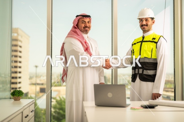 النظر إلى الكاميرا بتعابير السرور، الشركات المختصة بورشات البناء والمقاولات، تصميم وتخطيط المخططات المعمارية، رجل أعمال عربي خليجي سعودي يرتدي الشماغ والثوب التقليدي يقوم بمصافحة مهندس يرتدي الخوذة وسترة الحماية داخل المكتب، استخدام جهاز تقني حديث