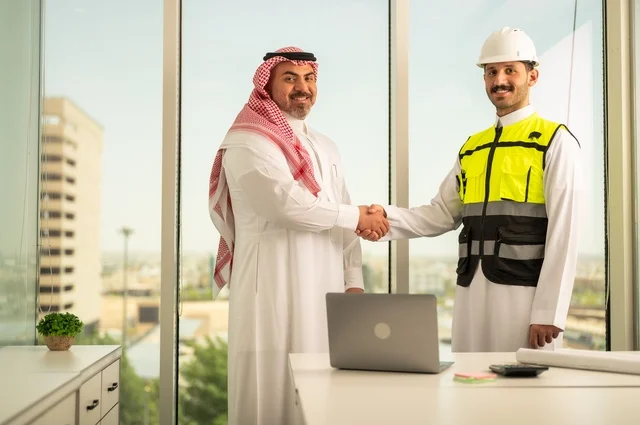 Looking at the camera with a happy expression, companies specialized in construction and contracting workshops, designing and planning architectural plans, a Saudi Arabian Gulf businessman wearing a traditional shemagh and thobe shaking hands with an engineer wearing a helmet and a protective jacket inside the office, using a modern technical device