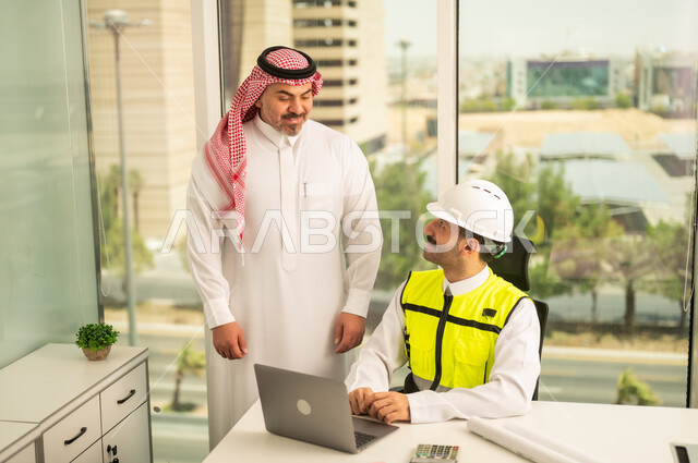 تصميم وتخطيط المخططات المعمارية عبر الحاسوب المحمول، متابعة تنفيذ الخطط الهندسية، الشركات المختصة بورشات البناء والمقاولات، رجل أعمال عربي خليجي سعودي يرتدي الشماغ ومهندس يرتدي الخوذة وسترة الحماية يقومان بالمناقشة وتبادل الخبرات، استخدام جهاز تقني حديث