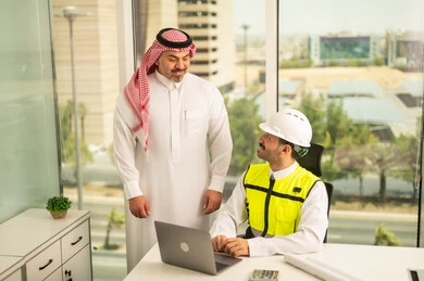 تصميم وتخطيط المخططات المعمارية عبر الحاسوب المحمول، متابعة تنفيذ الخطط الهندسية، الشركات المختصة بورشات البناء والمقاولات، رجل أعمال عربي خليجي سعودي يرتدي الشماغ ومهندس يرتدي الخوذة وسترة الحماية يقومان بالمناقشة وتبادل الخبرات، استخدام جهاز تقني حديث