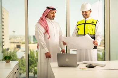 الشركات المختصة بورشات البناء والمقاولات، التخطيط ووضع اللمسات الأخيرة على المخططات المعمارية، رجل أعمال عربي خليجي سعودي يرتدي الشماغ ومهندس يرتدي الخوذة وسترة الحماية يعملان على مناقشة مخطط الإنشاء وتحديد القياسات بدقة، متابعة تنفيذ الخطط الهندسية الموضوعة مسبقا