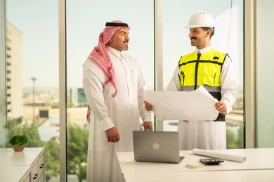 Following up on the implementation of pre-established engineering plans, companies specialized in construction and contracting workshops, planning and putting the final touches on architectural plans, a Saudi Arabian Gulf businessman wearing a shemagh and an engineer wearing a helmet and a protective vest working to discuss the construction plan and determine the precise measurements