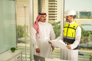 Companies specializing in construction and contracting workshops, planning and finalizing architectural plans. A Saudi Arabian Gulf businessman wearing a shemagh and an engineer wearing a helmet and a protective vest are working to discuss the construction plan and accurately determine measurements. They are following up on the implementation of the engineering plans drawn up in advance.