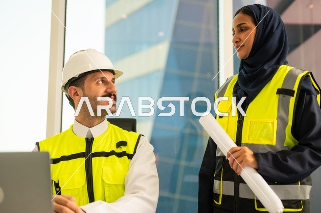 استخدام جهاز الحاسوب المحمول في انجاز الاعمال الهندسية،الوحدة والعمل الجماعي لتحقيق اهداف الشركه ، الشركات المختصة بورشات البناء والمقاولات، تصميم وتخطيط المخططات المعمارية،مهندس و مهندسة عربيتان خليجيان سعوديان يرتديان سترة وقاية يقومان بالعمل داخل مكتب هندسي