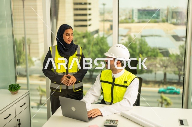 استخدام جهاز الحاسوب المحمول في انجاز الاعمال الهندسية،الوحدة والعمل الجماعي لتحقيق اهداف الشركه ، الشركات المختصة بورشات البناء والمقاولات، تصميم وتخطيط المخططات المعمارية،مهندس و مهندسة عربيتان خليجيان سعوديان يرتديان سترة وقاية يقومان بالعمل داخل مكتب هندسي