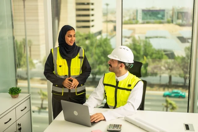 Using a laptop to accomplish engineering tasks, unity and teamwork to achieve the company's goals, companies specialized in construction workshops and contracting, designing and planning architectural plans, an Arab Gulf engineer and engineer from Saudi Arabia wearing protective vests working inside an engineering office.
