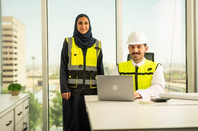 النظر الى الكاميرا بتعابير السرور ، استخدام جهاز الحاسوب المحمول في انجاز الاعمال الهندسية،الوحدة والعمل الجماعي لتحقيق اهداف الشركه ، الشركات المختصة بورشات البناء والمقاولات، تصميم وتخطيط المخططات المعمارية،مهندس و مهندسة عربيتان خليجيان سعوديان يرتديان سترة وقاية