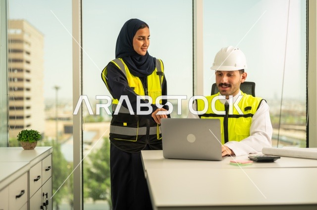 استخدام جهاز الحاسوب المحمول في انجاز الاعمال الهندسية،الوحدة والعمل الجماعي لتحقيق اهداف الشركه ، الشركات المختصة بورشات البناء والمقاولات، تصميم وتخطيط المخططات المعمارية،مهندس و مهندسة عربيتان خليجيان سعوديان يرتديان سترة وقاية يقومان بالعمل داخل مكتب هندسي