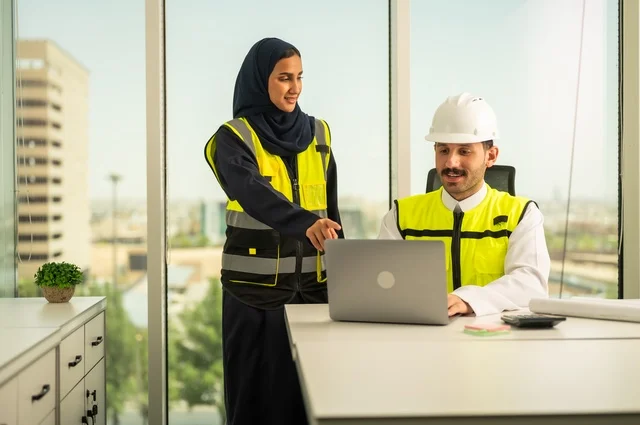 استخدام جهاز الحاسوب المحمول في انجاز الاعمال الهندسية،الوحدة والعمل الجماعي لتحقيق اهداف الشركه ، الشركات المختصة بورشات البناء والمقاولات، تصميم وتخطيط المخططات المعمارية،مهندس و مهندسة عربيتان خليجيان سعوديان يرتديان سترة وقاية يقومان بالعمل داخل مكتب هندسي