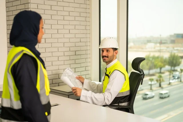 Completion of engineering works, unity and teamwork to achieve the company's goals, companies specialized in construction workshops and contracting, design and planning of architectural plans, an Arab Gulf engineer and engineer from Saudi Arabia wearing safety vests are discussing and exchanging experiences.