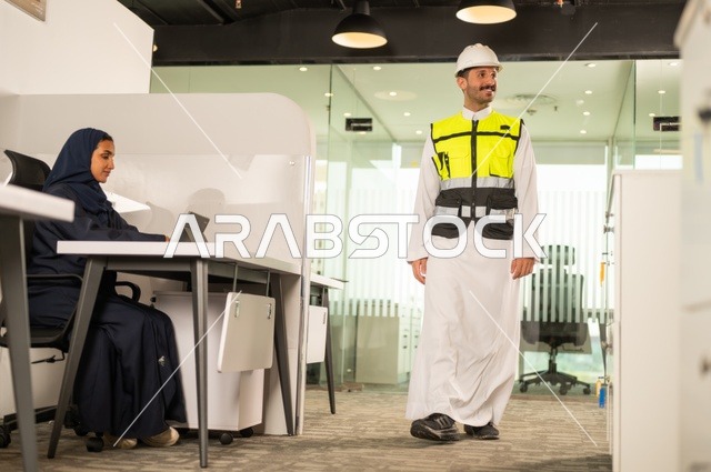 تصميم وتخطيط المخططات المعمارية،انجاز الاعمال الهندسية، الشركات المختصة بورشات البناء والمقاولات، مهندس عربي خليجي سعودي يرتدي السترة و خوذوة الحماية يقوم بالتجول داخل الشركة ، موظفة محجبة تجلس خلف المكتب تقوم بالعمل عبر الجهاز اللوحي