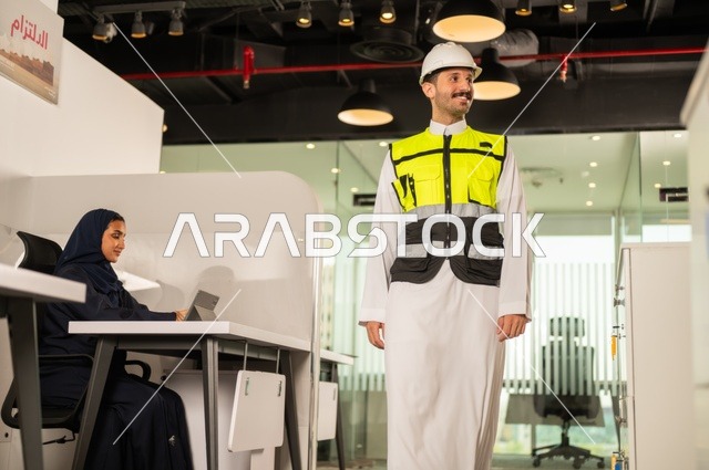 تصميم وتخطيط المخططات المعمارية،انجاز الاعمال الهندسية، الشركات المختصة بورشات البناء والمقاولات، مهندس عربي خليجي سعودي يرتدي السترة و خوذوة الحماية يقوم بالتجول داخل الشركة ، موظفة محجبة تجلس خلف المكتب تقوم بالعمل عبر الجهاز اللوحي