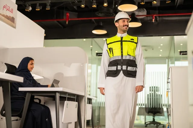 تصميم وتخطيط المخططات المعمارية،انجاز الاعمال الهندسية، الشركات المختصة بورشات البناء والمقاولات، مهندس عربي خليجي سعودي يرتدي السترة و خوذوة الحماية يقوم بالتجول داخل الشركة ، موظفة محجبة تجلس خلف المكتب تقوم بالعمل عبر الجهاز اللوحي