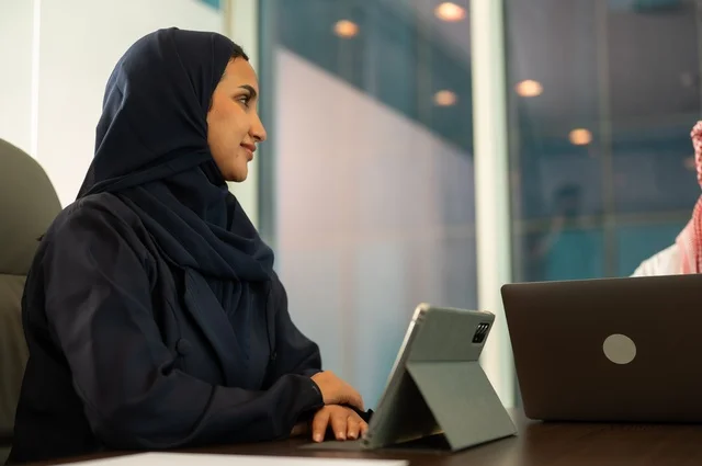 التوظيف في المكاتب والشركات السعودية ،مهن وأعمال مكتبية، توفير بيئة عمل مريحة ومناسبة، استخدام جهاز الحاسوب المحمول لإنجاز المهام باحترافية، سيدة عربية خليجية سعودية ترتدي العباءة تجلس في المكتب تقوم بالعمل عبر الجهاز اللوحي