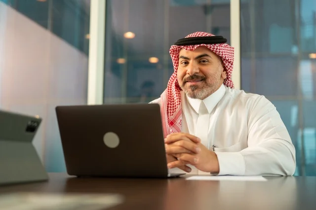 مهن وأعمال مكتبية، إيماءات الاندماج والتركيز، توفير بيئة عمل مريحة ومناسبة، استخدام جهاز الحاسوب المحمول لإنجاز المهام باحترافية، رجل عربي خليجي سعودي يرتدي الثوب والشماغ التقليدي يجلس في المكتب يقوم بالعمل عبر اللابتوب، التوظيف في المكاتب والشركات السعودية