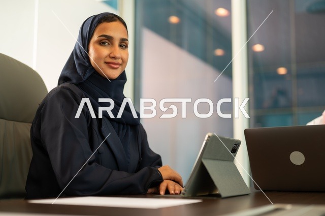 التوظيف في المكاتب والشركات السعودية ،مهن وأعمال مكتبية، توفير بيئة عمل مريحة ومناسبة، استخدام جهاز الحاسوب المحمول لإنجاز المهام باحترافية، سيدة عربية خليجية سعودية ترتدي العباءة تجلس في المكتب تقوم بالعمل عبر الجهاز اللوحي