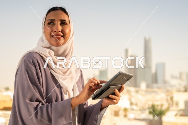 التواصل مع الاهل و الاصدقاء ،جهاز تقني حديث و متطور،تصفح الانترنت و مواقع التواصل الاجتماعي عبر جهاز التابليت ،صورة  مقربة لشابة عربية خليجية سعودية ترتدي العباءة تقوم باستخدام الجهاز اللوحي بايماءات السعادة والسرور،الخروج في الهواء الطلق
