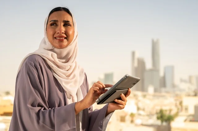Communicating with family and friends, a modern and advanced technical device, browsing the internet and social media sites via a tablet, a close-up image of a young Arab Gulf Saudi woman wearing an abaya using the tablet with gestures of happiness and joy, going outdoors.