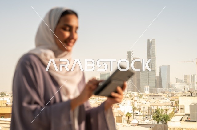 التواصل مع الاهل و الاصدقاء ،جهاز تقني حديث و متطور،تصفح الانترنت و مواقع التواصل الاجتماعي عبر جهاز التابليت ،صورة  مقربة لشابة عربية خليجية سعودية ترتدي العباءة تقوم باستخدام الجهاز اللوحي بايماءات السعادة والسرور،الخروج في الهواء الطلق