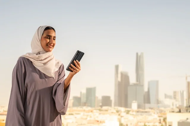 Browsing the internet and social media through a mobile device, an Arab Gulf Saudi woman wearing an abaya is using her mobile phone with gestures of happiness and joy, going outdoors, and communicating with family and friends.