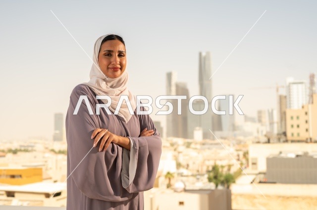 مفهوم الاناقة والاهتمام بالمظهر الخارجي، شابة عربية خليجية سعودية ترتدي العباءة تقف بايدي متقاطعة ، النظر الى شيء ما بايماءات السعادة والسرور،الخروج في الهواء الطلق