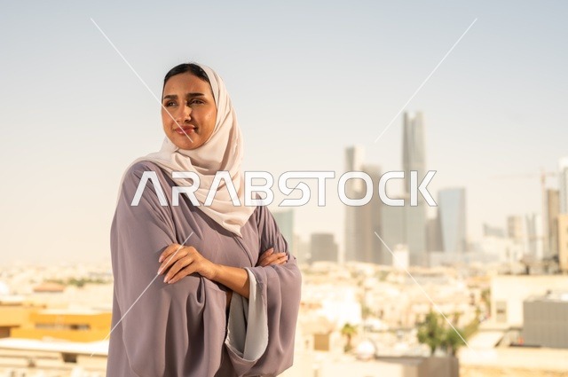مفهوم الاناقة والاهتمام بالمظهر الخارجي، شابة عربية خليجية سعودية ترتدي العباءة تقف بايدي متقاطعة ، النظر الى شيء ما بايماءات السعادة والسرور،الخروج في الهواء الطلق