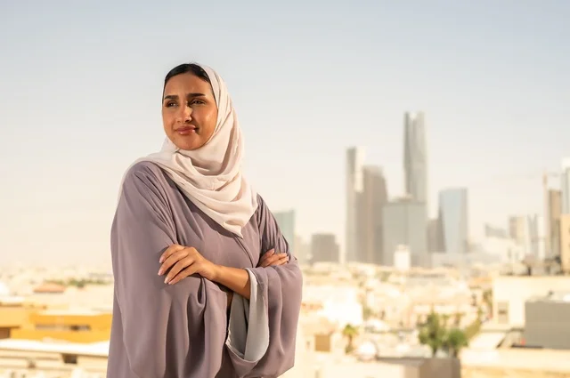 The concept of elegance and attention to appearance, a young Arab Gulf Saudi woman wearing an abaya stands with her arms crossed, looking at something with gestures of happiness and joy, going out in the open air.